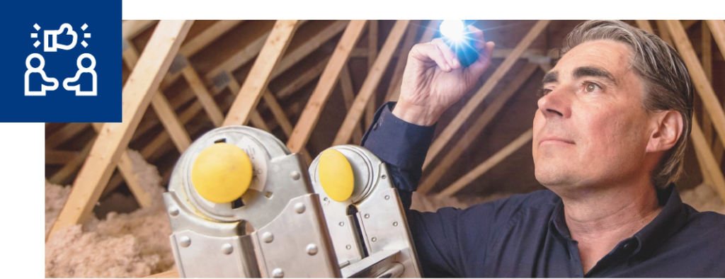 A home inspector uses a flash light to inspect the attic while he stands on a ladder. In the top left hand corner there is a blue box with a white icon of two people shaking hands and a thumbs up above them.