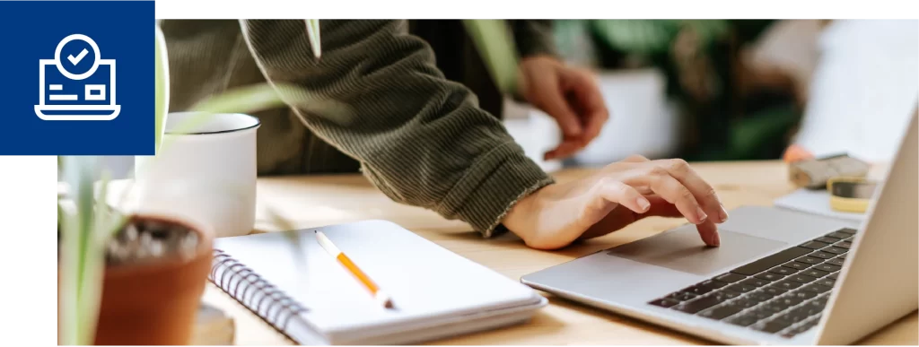 Hand using a laptop trackpad on a desk next to a spiral notebook and mug.