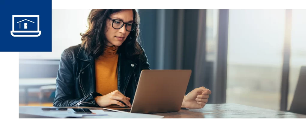 Woman in glasses working on a laptop at a table in a bright room.