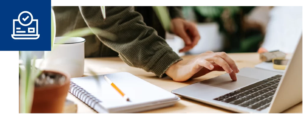 Close-up of a hand using a laptop on a desk beside a notebook and mug.