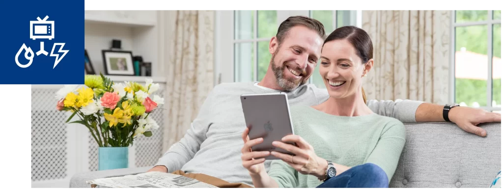 Smiling couple sitting on a couch at home looking at a tablet together, with flowers on a nearby table.